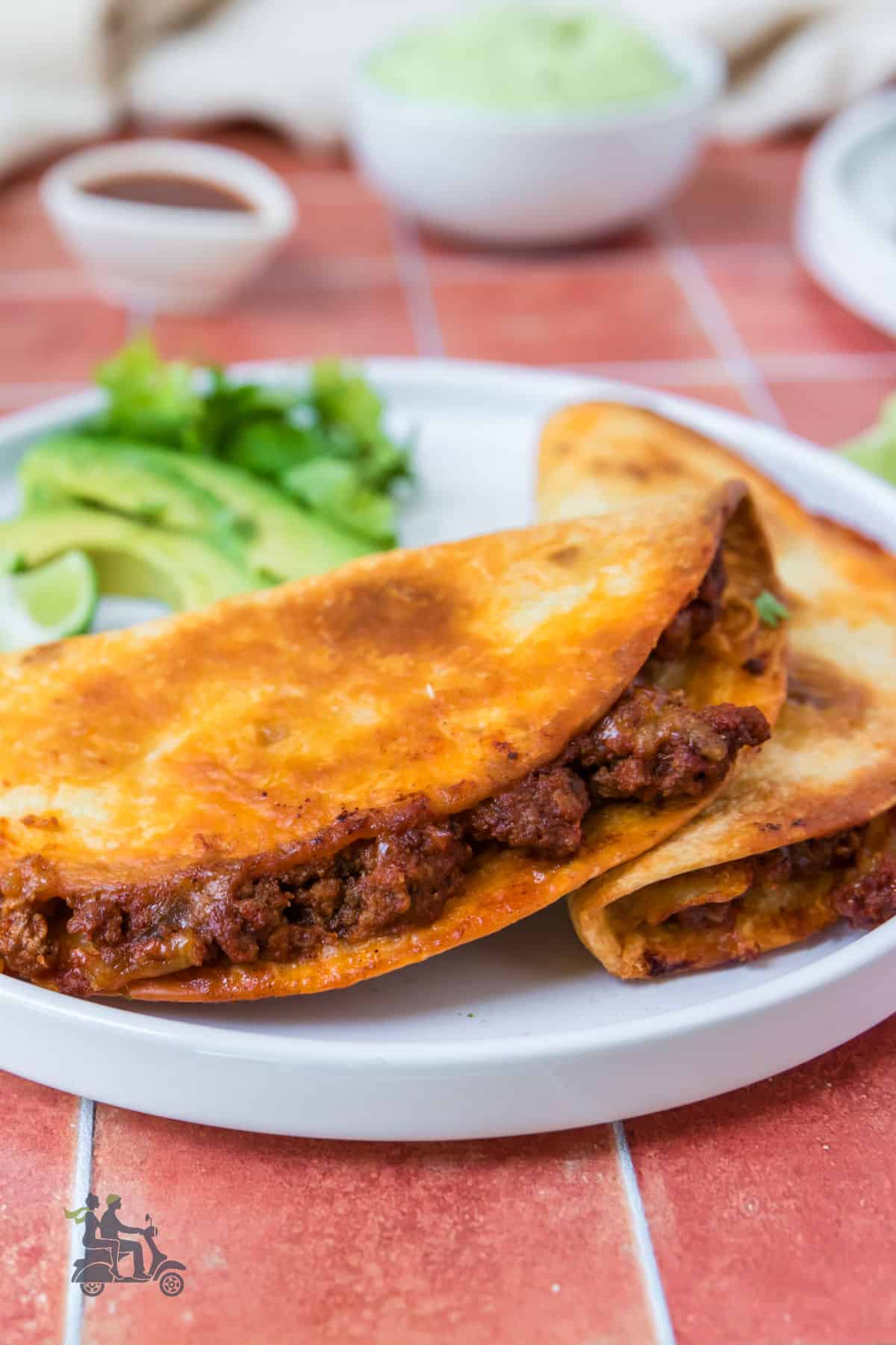 Two crispy beef tacos on a white plate with avocado slices on the side.