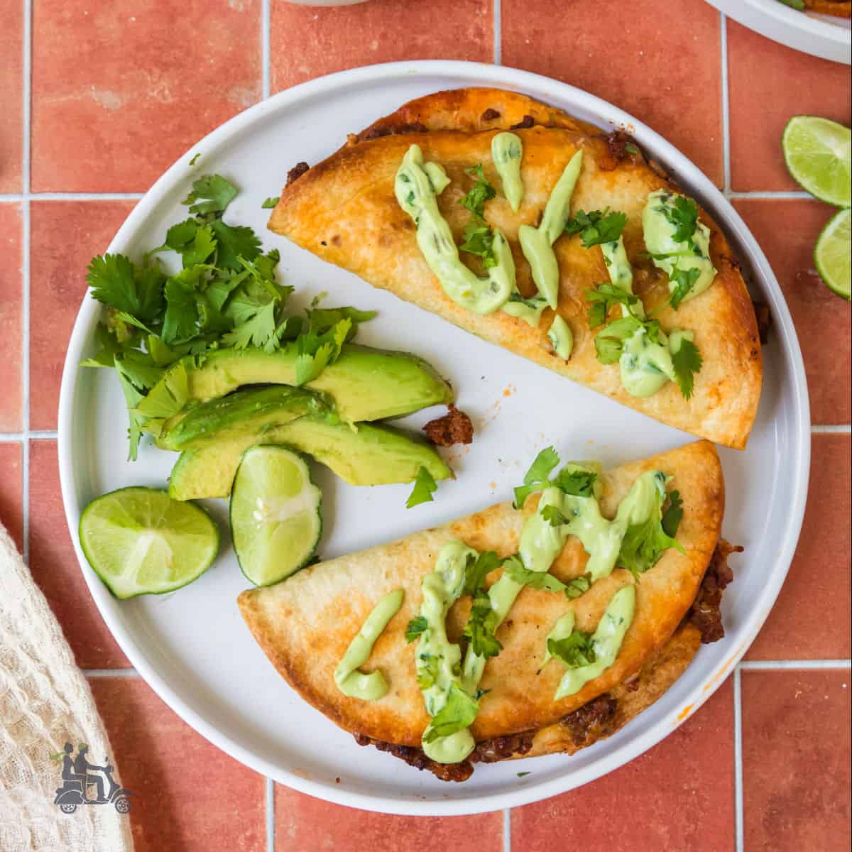 Two oven-baked beef tacos on a white plate with lime wedges, avocado slices, and chopped cilantro.
