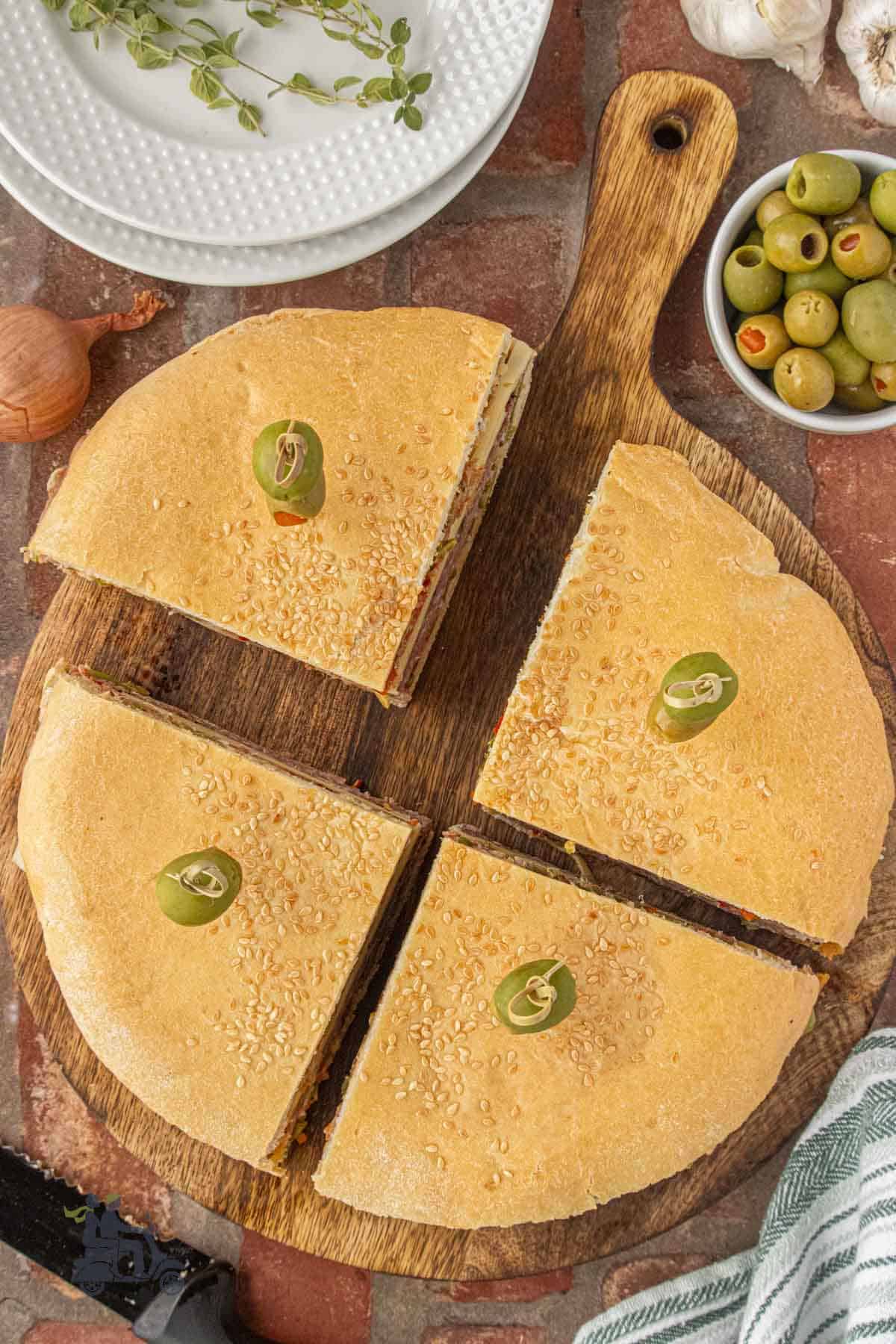 A muffuletta loaf sits on a cutting board sliced into four wedges each with an olive garnish.