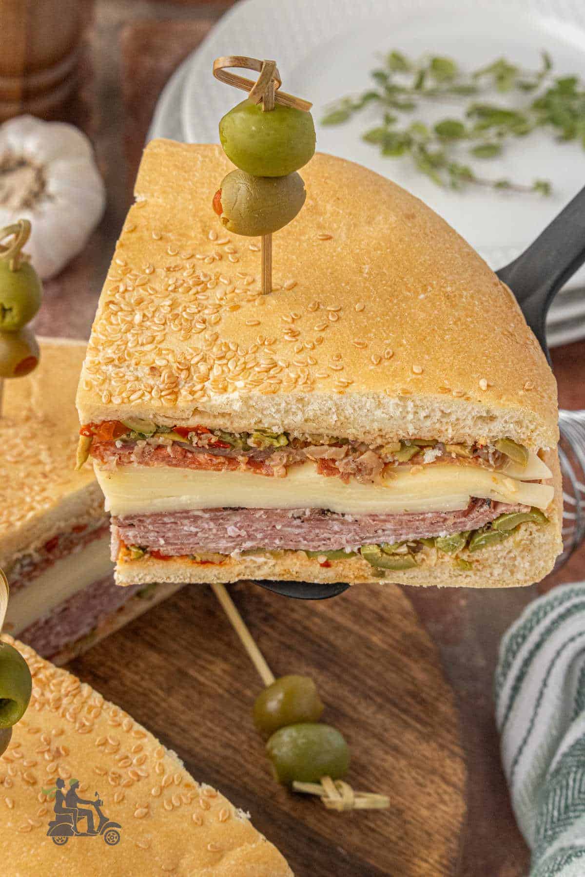 A spatula scoops up a muffuletta sandwich wedge with a garnish of olives on a toothpick.