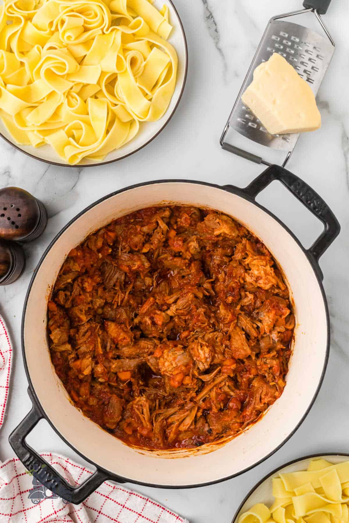 Here's a revised version of your text:
"An overhead view shows a Dutch oven filled with pork bolognese sauce. Nearby, plates hold pappardelle pasta, ready for the sauce."