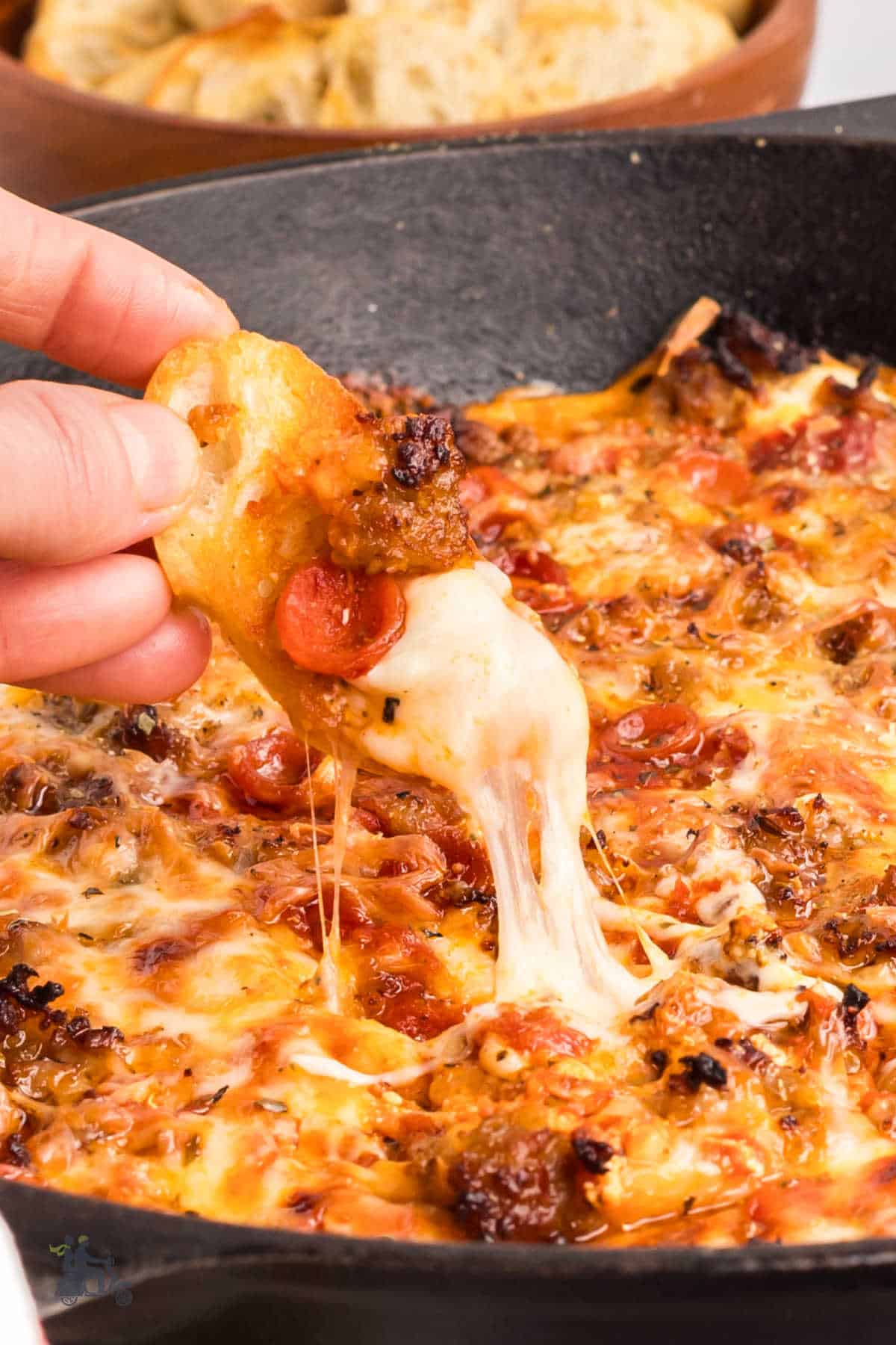 Hot pizza dip with sausage and pepperoni in a black skillet with a crostini dipper dipping a scoop.