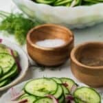 A long pin showing a summer cucumber salad served as a side dish with salt and black pepper on the side.