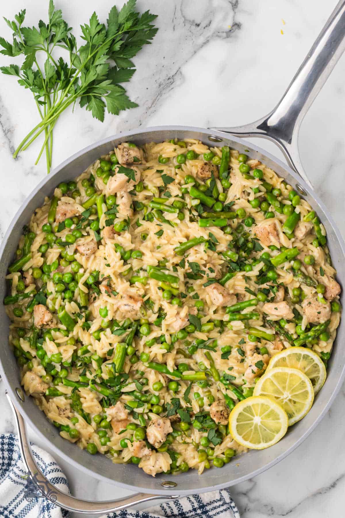 Creamy lemon chicken orzo primavera with asparagus and peas in a skillet with lemon slices as a garnish.