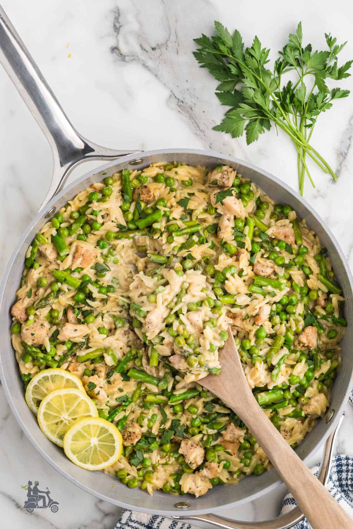 Wooden spoon scoops up some of the bright and creamy chicken orzo primavera pasta dish from the skillet.