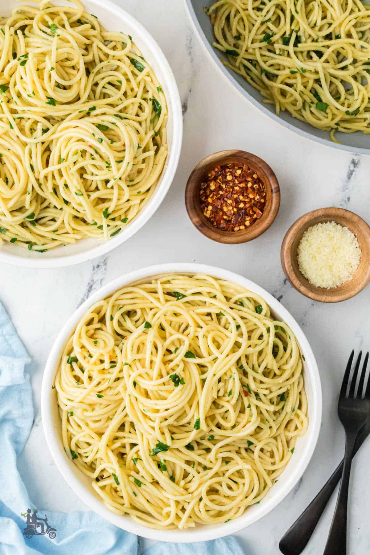 Overhead view of two bowls of spaghetti aglio e olio. The pasta is tossed with olive oil, garlic, and peperoncino. It is finished with flat-leaf parsley and grated Parmesan.