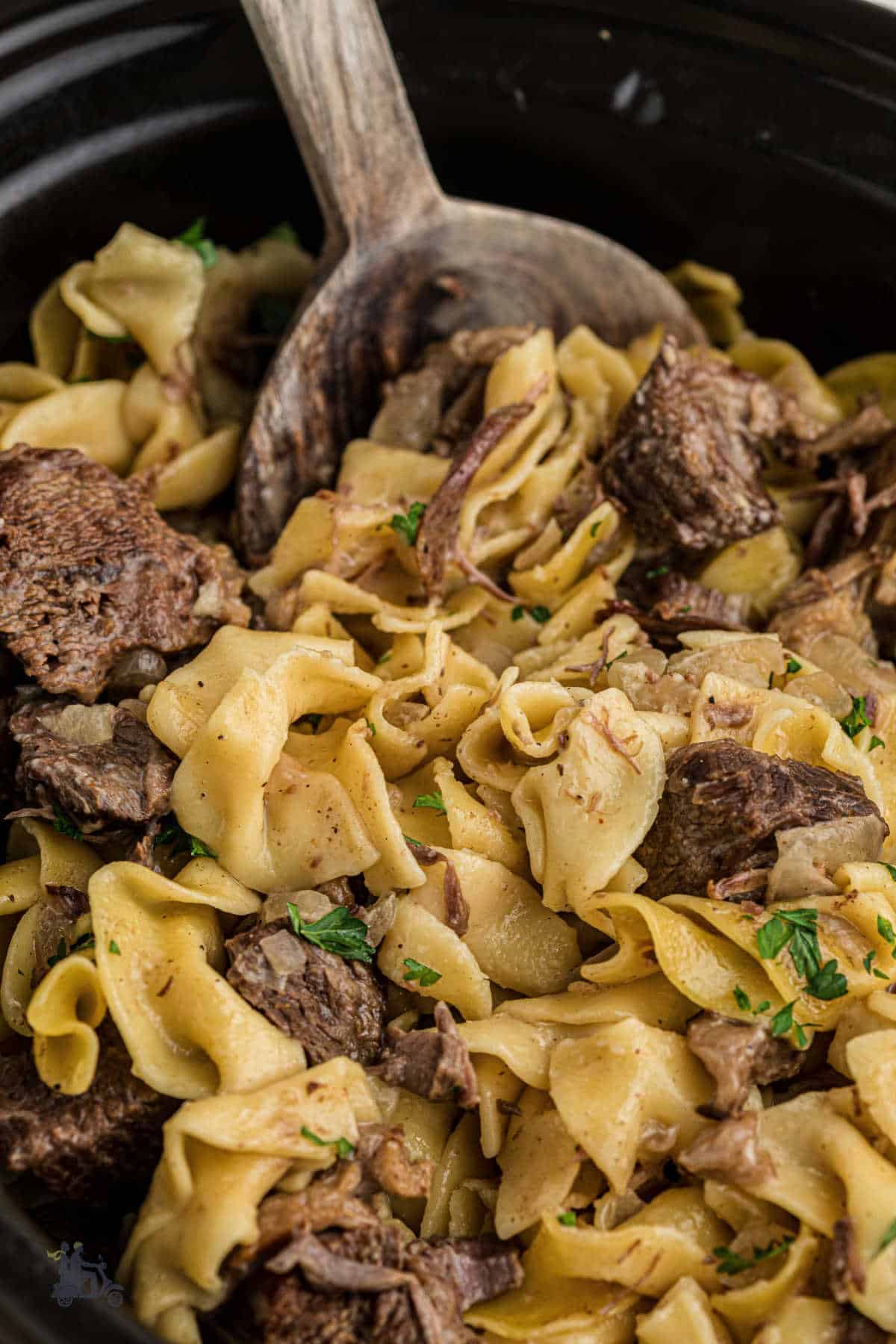 A wooden spoon scoops up a large helping of wide egg noodles and tender cubes of seasoned chuck roast.